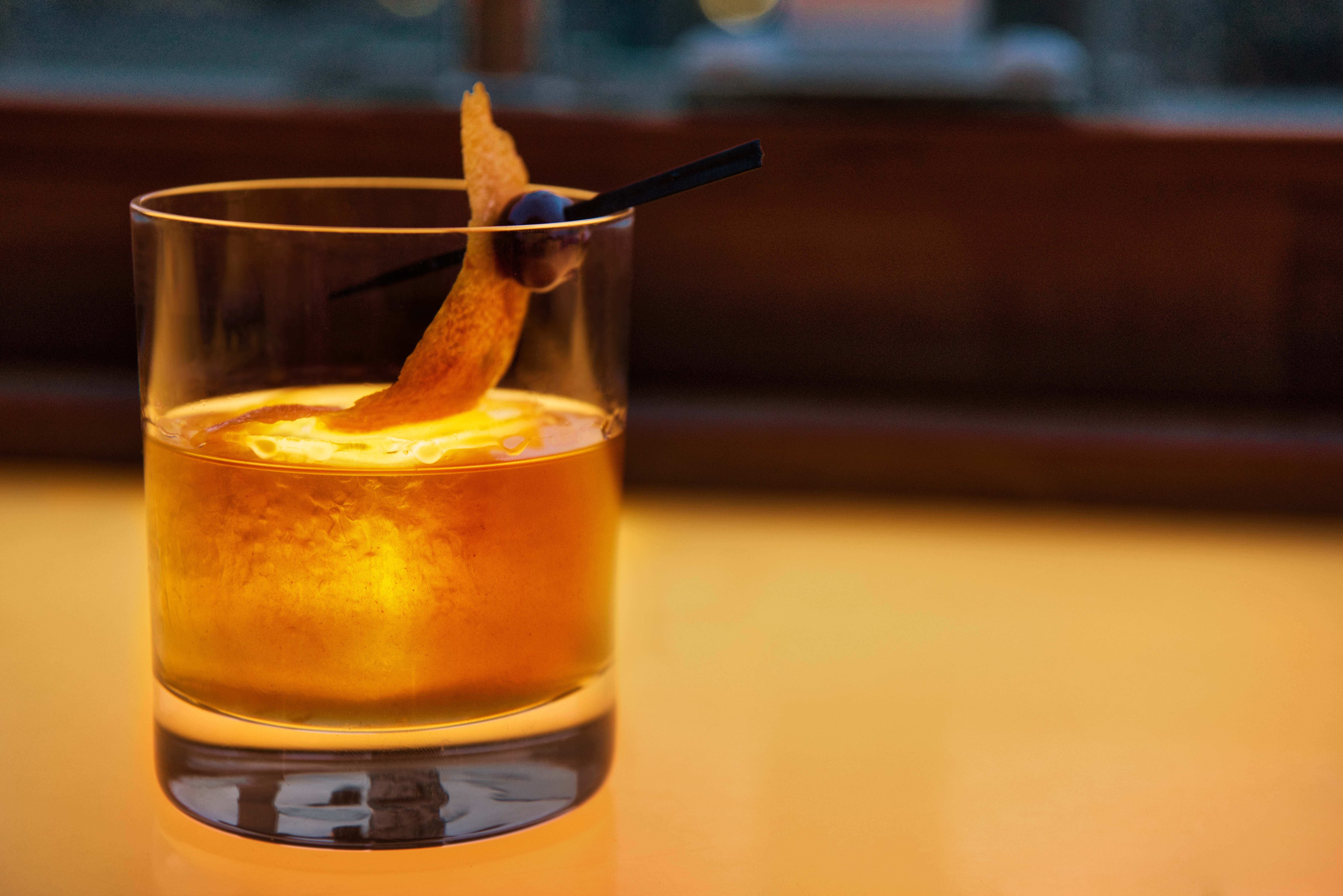 Bright orange Old Fashioned cocktail in a glass with orange peel and cherry garnish